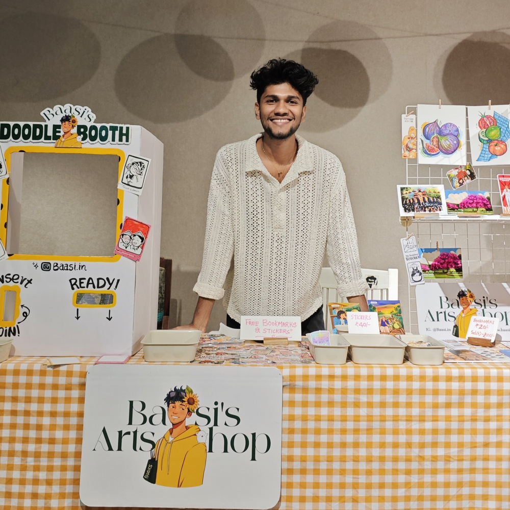 Baasi standing behind the live doodle booth at a Bengaluru event, with the yellow gingham table, READY! window and doodle cards